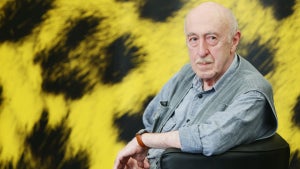 An older light-skinned man sits, turning toward the camera in a chair, in front of a yellow fur background with black spots.