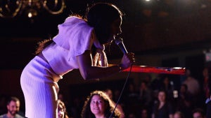 A Black woman stands on stage in a pink top and skirt, holding a microphone, in front of an audience.
