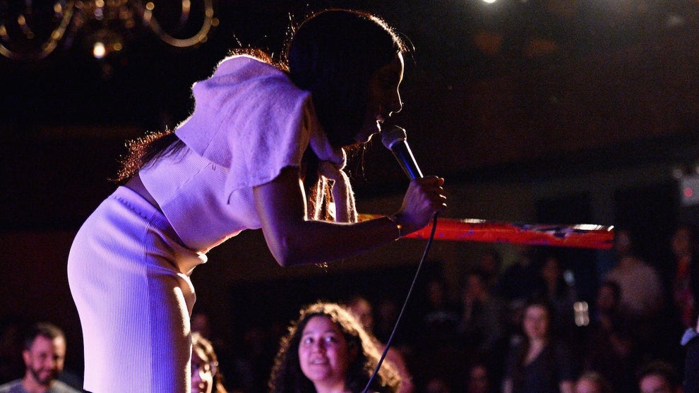 A Black woman stands on stage in a pink top and skirt, holding a microphone, in front of an audience.