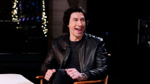 A man with light-toned skin wearing a leather jacket sitting in a director's chair on a dark stage.