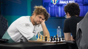 A man in a white shirt leans on his arm as he plays chess against a man with a black shirt with his back to the camera.