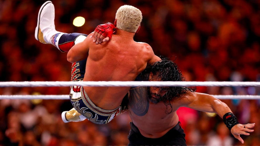 Two pro wrestlers wrestle, one with medium-light-toned skin and the other with medium-toned skin. They are in the middle of a move in a ring with a larger crowd seen sitting in the background.
