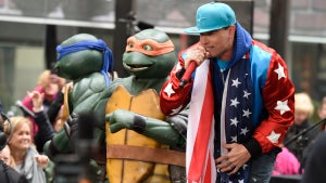 Two humanoid turtles in ninja costumes on stage in front of an outdoor crowd with a light-skinned man wearing an American flag around his neck, a light blue ball cap, and an American-themed jacket featuring white stars on red satin-sheen sleeves.