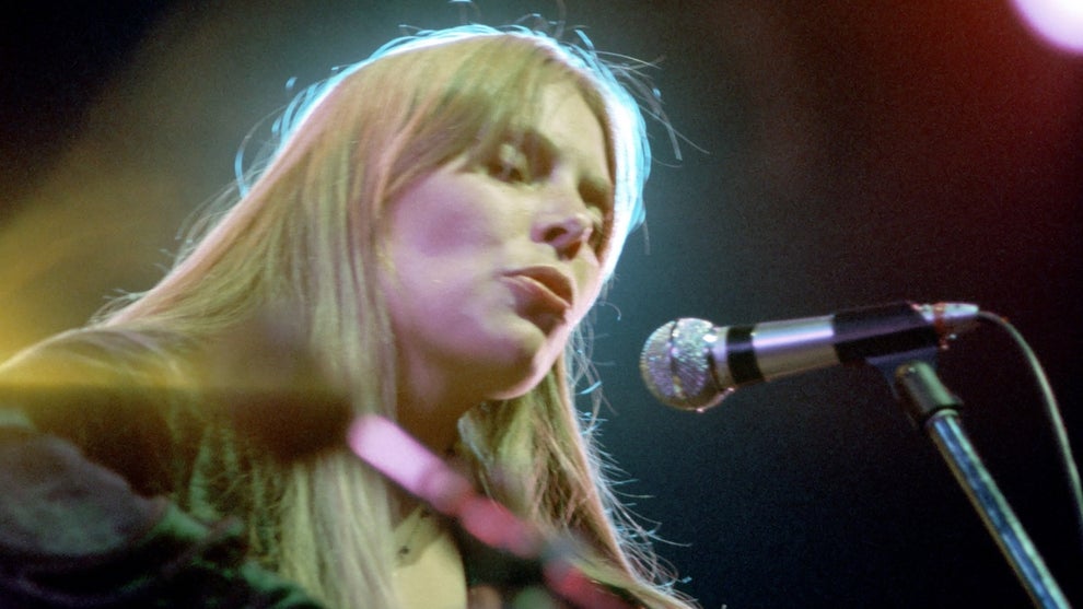 A woman with light-toned skin and blond hair performs, eyes closed, in front of a microphone stand, with a halo of light around her head. She appears to be playing a guitar. The woman is Joni Mitchell.