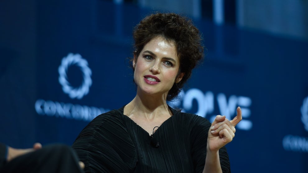 A woman with light-toned skin gestures thoughtfully on a stage with a medium-blue backdrop and logos.