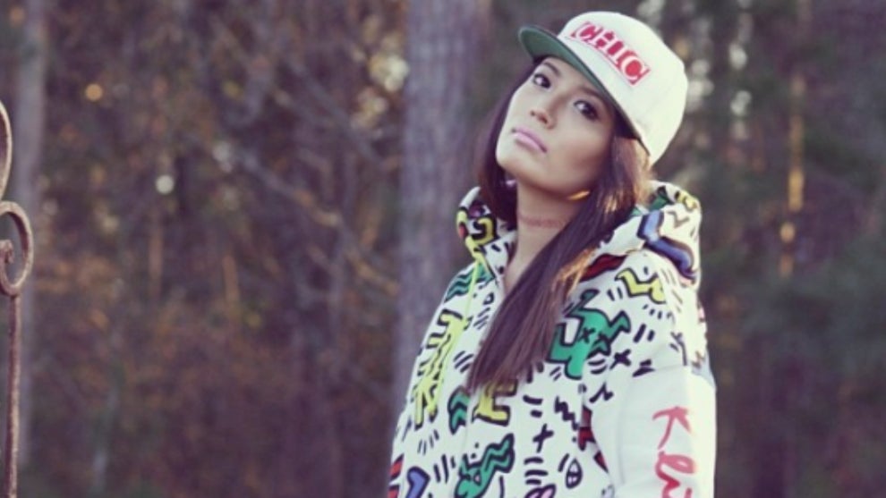 A woman with medium-toned skin poses in a bright hoodie and baseball cap in a forest