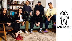 A group of people sit and stand around a room, including on a couch, with bookshelves in the background. On the right is a logo labeled "MUTANT" showing a one-eyed alien figure with arms and legs, a bit reminiscent of the characters from Among Us.