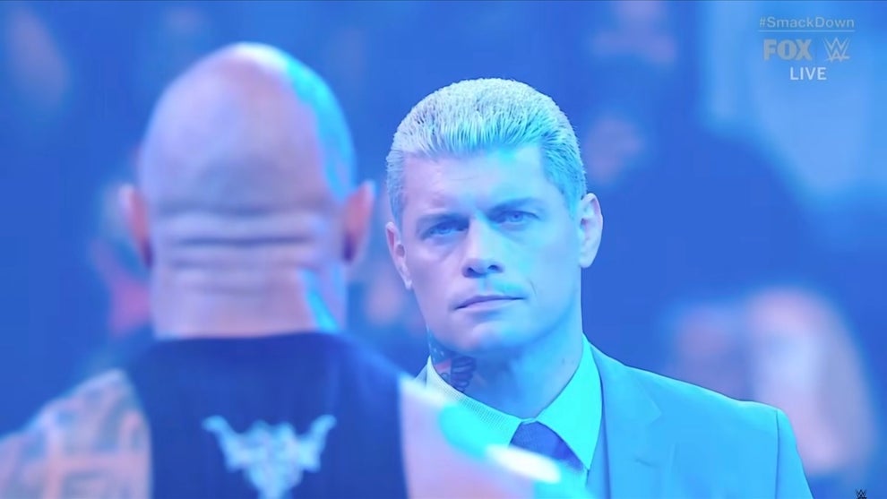 A man with medium-toned skin is seen from behind, facing off with a man with light-toned skin. They are Dwayne "The Rock" Johnson and Cody Rhodes, pro wrestlers, standing in a wreslting ring in front of a crowd.