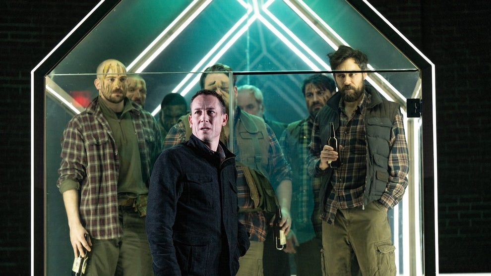 A closeup of actors crowded inside a glass house on stage, lit up with stage lighting.