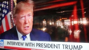 A television shows Donald Trump sitting down for an interview in a room with wood paneling and an American flag behind him. He has light-toned skin with a reddish hue. Text at the bottom of the screen shows that it's from Fox News and reads "INTERVIEW WITH PRESIDENT TRUMP."