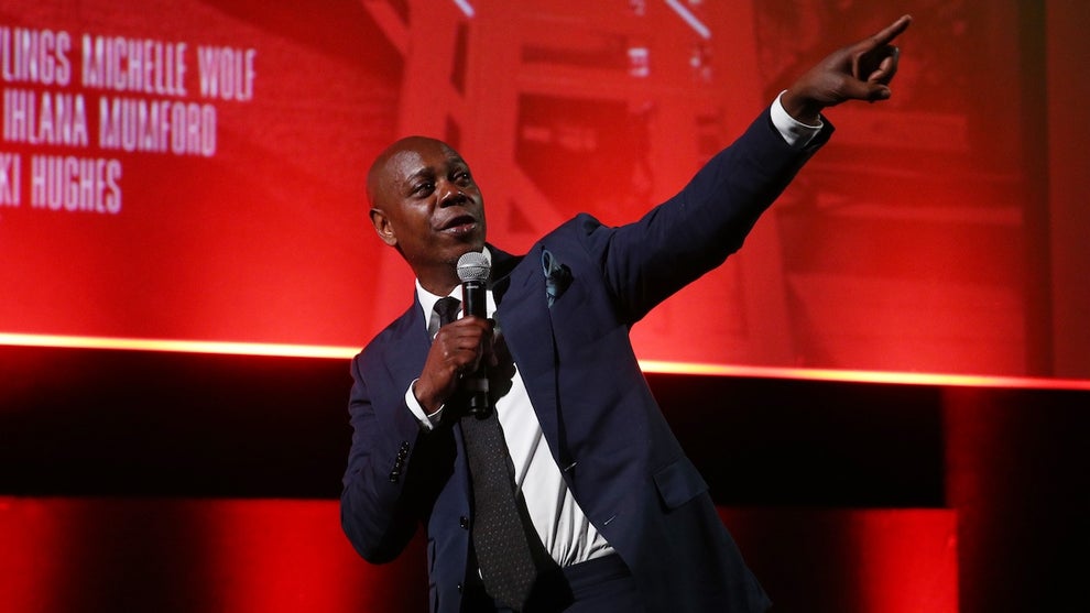 A dark-skinned man wearing a suit stands on stage, pointing up and to the right, in front of a red cinema projection background. The man is comedian Dave Chappelle.