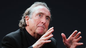 An older white man looks slightly upward as he gestures with both hands, a far-off look in his eyes. He wears a suit and has graying hair.