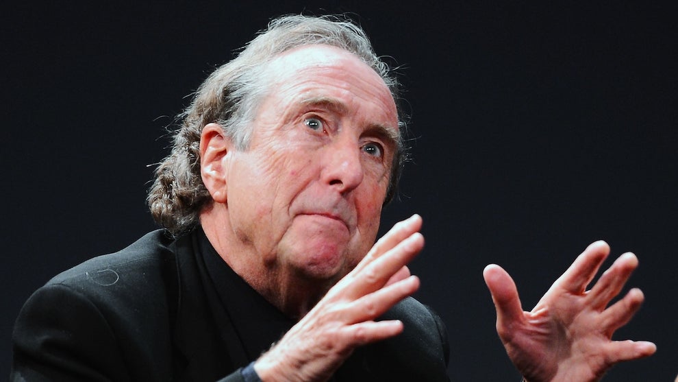 An older white man looks slightly upward as he gestures with both hands, a far-off look in his eyes. He wears a suit and has graying hair.