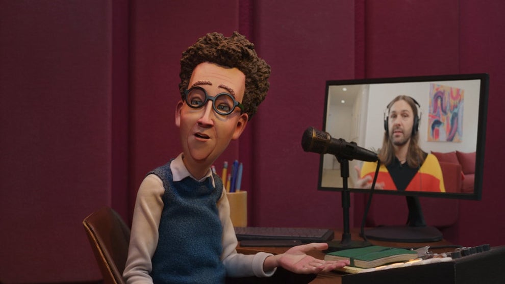 A puppet of a middle-aged white man on the left with a video screen showing an image of a middle-aged white man with long hair and a colorful sweater. The puppet man sits in front of a microphone and control board.
