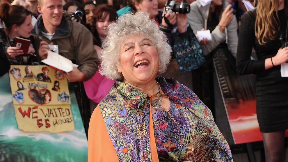 A woman stands smiling, wearing a cape-like outfit, on the red carpet.