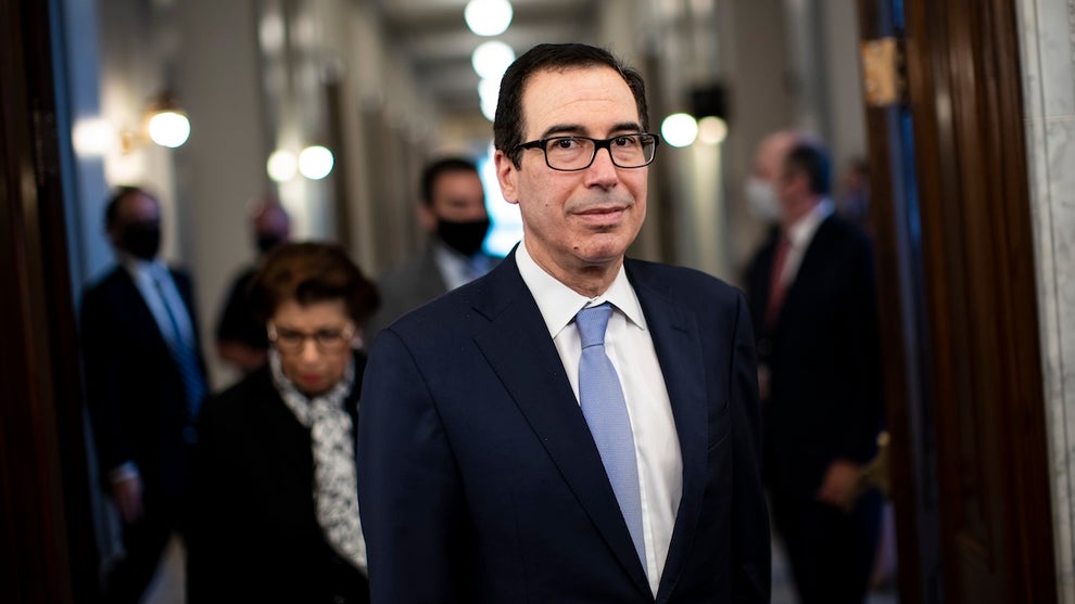Steven Mnuchin, a man with light-toned skin, looks at the camera as he stands in front of a hallway with others behind him. He wears a suit.