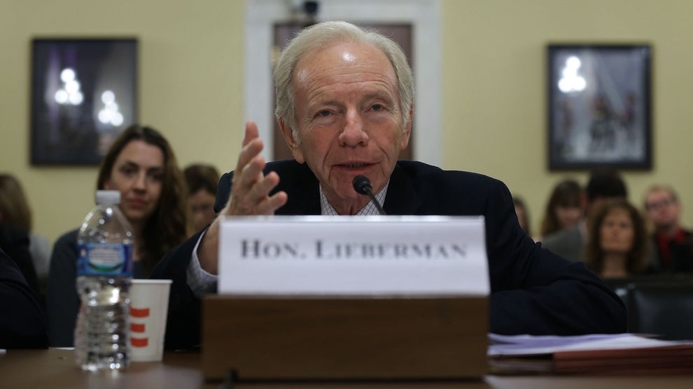 An older man with light-toned skin speaks sitting at a table with a nameplate reading "HON. LIEBERMAN"