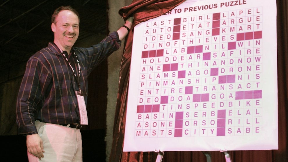 A man with a mustache stands smiling, pulling a covering over a large board revealing the answer to a crossword puzzle. The man has light-toned skin and appears middle aged.