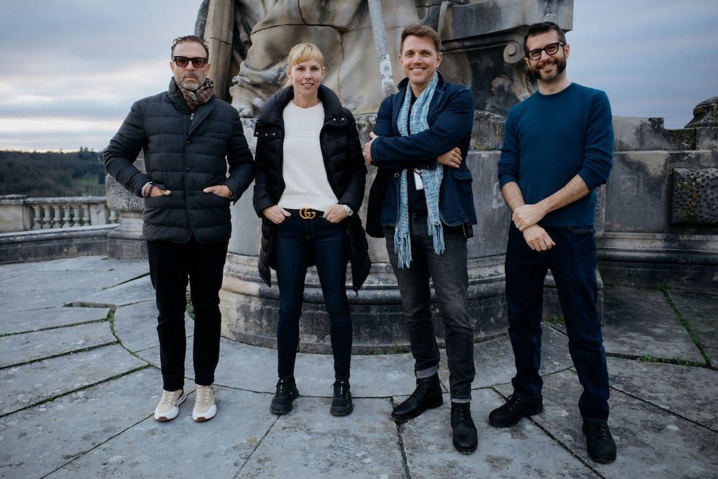 Four people stand outside in front of a stone statue. They are four people with light-toned skin, including three men and one woman.