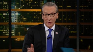 A man with light-toned skin makes a confused face on the set of a talk show, holding a blue notecard in one hand.