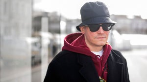 A man with light-toned skin wearing a bucket hat and sunglasses with a hoodie and a jacket, standing outside.
