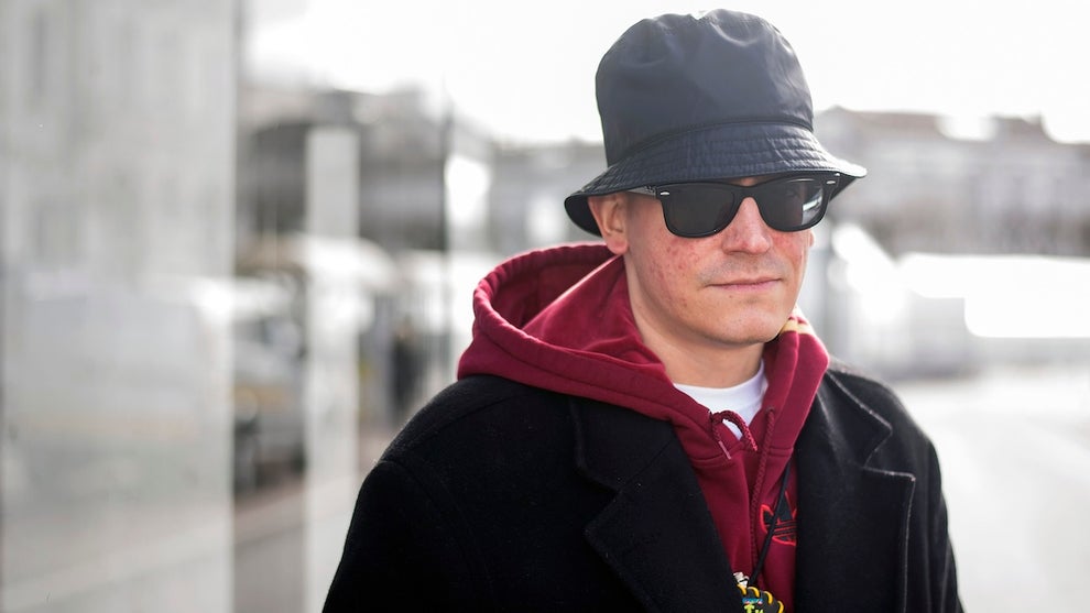A man with light-toned skin wearing a bucket hat and sunglasses with a hoodie and a jacket, standing outside.