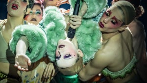 A woman in a mint green fur coat lays holding a microphone in the arms of dancers, their head upside-down as they reach out. She wears near-white makeup.