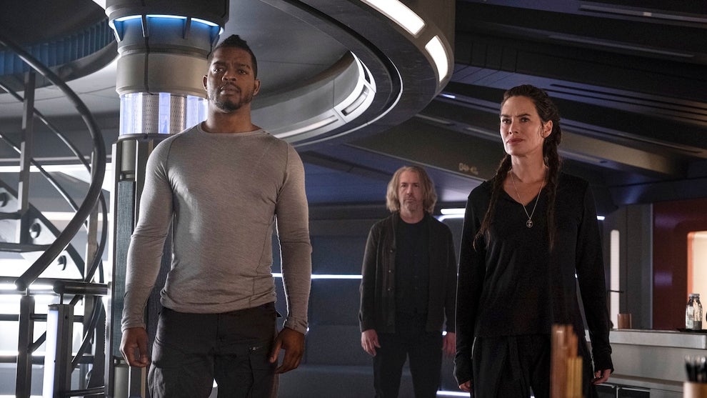 Three people stand inside a sci-fi setting, a space beacon. They include a Black man, a white man, and a white woman. The white man stands in the center, behind the other two. They all are looking toward something off-camera, apparently in awe.
