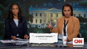 Two Black women wearing blazers sit at an anchor table with an image of the White House behind them. A quote on the screen shows an excerpt reading "a trained assassin."
