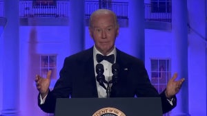 An older man, President Joe Biden, holds up his hands and raises his eyebrows with a somewhat disbelieving look on his face. He wears a tuxedo and stands in front of a backdrop showing the White House.