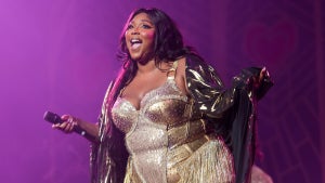 A Black woman smiles on stage, wearing a gold outfit with a light translucent shawl on top. She holds a microphone and stands on stage in front of a purple background.
