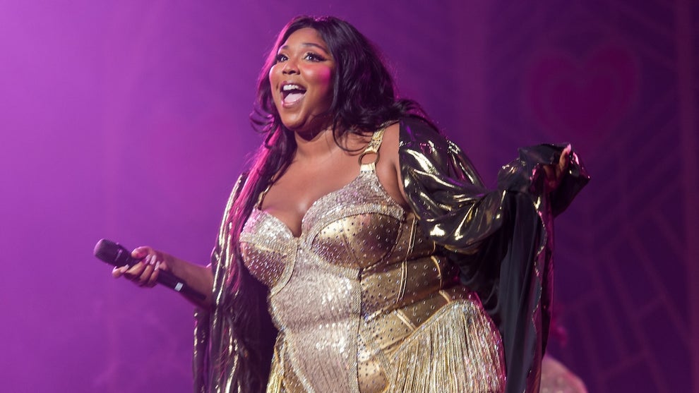 A Black woman smiles on stage, wearing a gold outfit with a light translucent shawl on top. She holds a microphone and stands on stage in front of a purple background.