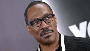 A closeup of a Black man's face. He wears glasses and has a mustache and close-cropped haircut. The man is Eddie Murphy, wearing a black jacket and standing in front of a gray background.