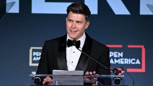A light-skinned man stands at a podium, smiling and waring a tuxedo.