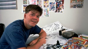 A young man with light-toned skin leans on an artist's table, featuring black and white sketches and an issue of the comic book "Youngblood." He has floppy hair. Behind him are more illustrations on the wall, including of his character Prophet in a closeup, as well as a Calvin and Hobbes comic strip pinned to the wall.