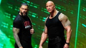 Two men with medium-toned skin with large muscles stand on stage with a bright green background. They both look perturbed. One has a title belt over his shoulder.