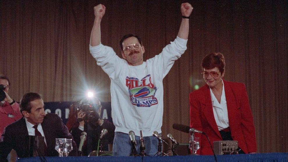 A man in the center stands triumphantly in front of microphones, arms in the air. He and the others in the photo have light-toned skin. He wears a white sweater with the sleeves rolled up.