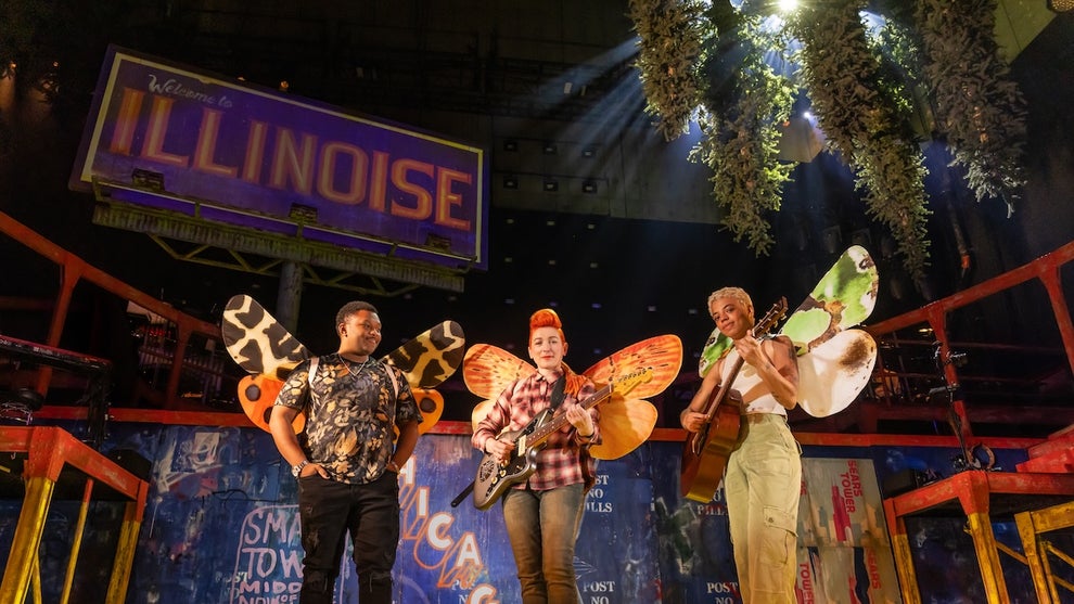 Three people stand holding guitars and dressed as moths, wearing large wings. A billboard in the background reads "Welcome to Illinoise." Light shines through a tree's branches in the upper right of the image.