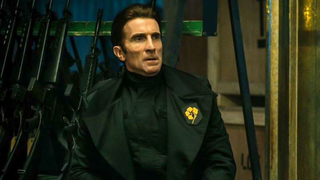 A man dresseded in an authoritarian-looking but stylish black suit with a yellow flower on the lapel stares at something out of frame