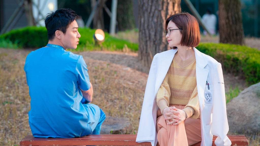 A man in scrubs sits on a bench with his back to the camera with a woman in a doctor's coat sits next to him, facing forward and smiling at him