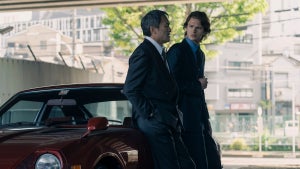 An actor with medium-toned skin and another with light-toned skin lean against a deep red sports car. The man on the left has a far-off expression while the man on the right looks surprised. The people are actors Ken Watanabe and Ansel Elgort from "Tokyo Vice."