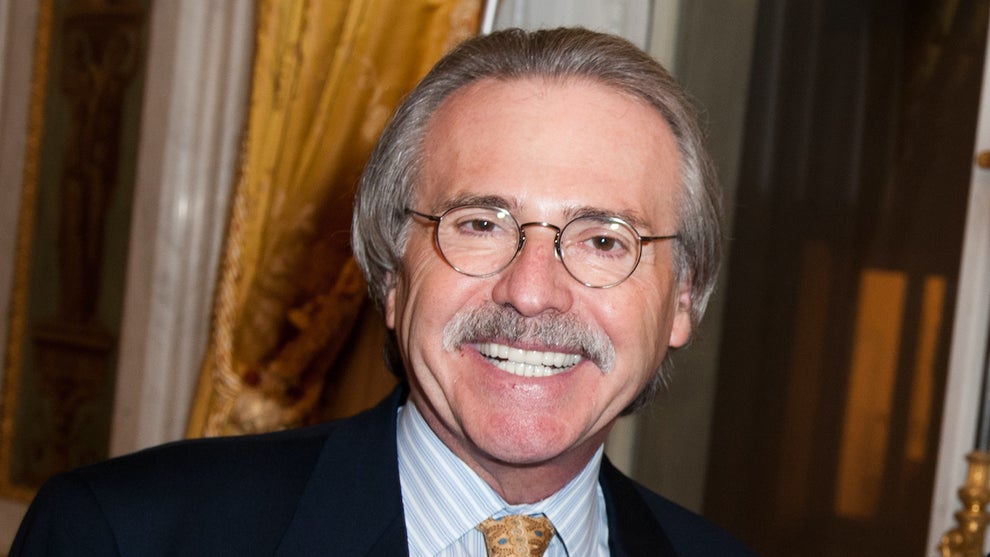 An older man with a silver mustache smiles, wearing a suit, in a ballroom-styled environment.