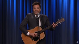 A man with light-toned skin (but with a tan) holds an acoustic guitar and wears a suit, performing in front of a mic stand, with a blue curtain behind him. The man is Jimmy Fallon.