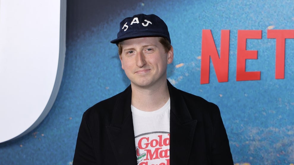 A man gives a small smile standing in front of a Netflix backdrop in light blue. He wears a hat that reads "JAJ." The man is James Austin Johnson.