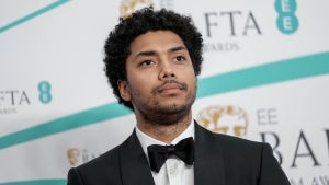 A man with dark-toned skin stands in front of a step-and-repeat background, wearing a tuxedo, with a solemn look on his face.