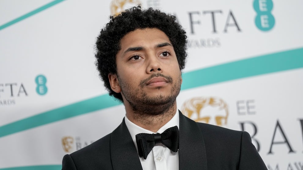 A man with dark-toned skin stands in front of a step-and-repeat background, wearing a tuxedo, with a solemn look on his face.