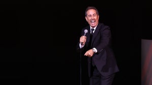 A man smiles and gestures as he holds a mic on stage. The light-skinned man wears a suit.