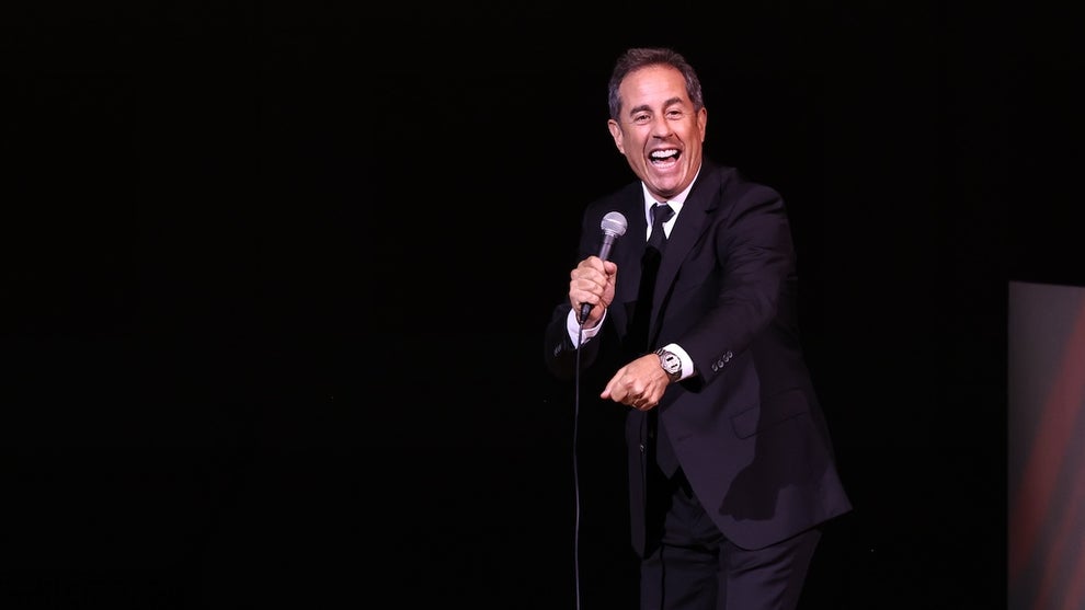 A man smiles and gestures as he holds a mic on stage. The light-skinned man wears a suit.