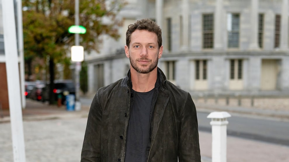 A man stands wearing a jacket on a city street, with a stone building and trees behind him.