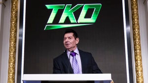 An older man with dyed hair and a mustache stands at a podium with the letters "TKO" behind him in a stylized green logo. The man is Vince McMahon.